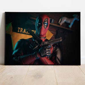 Puede incluir: Impresi&oacute;n enmarcada de Deadpool con un traje rojo y negro, sosteniendo dos pistolas negras. La m&aacute;scara del personaje es visible, con cubiertas negras para los ojos. El fondo incluye una pared de ladrillos y un letrero amarillo con la palabra "TRA".