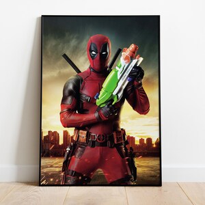 Puede incluir: Impresi&oacute;n enmarcada de Deadpool con su traje rojo y negro, sosteniendo una pistola de agua verde y blanca. El fondo muestra una silueta de paisaje urbano contra un cielo de atardecer. La impresi&oacute;n est&aacute; en un marco negro.