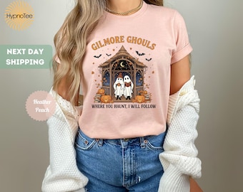 Może przedstawiać: Jasnoróżowa koszulka z motywem Halloween. Projekt zawiera napis "GILMORE GHOULS" nad ilustracją dwóch duchów w altanie, dyniami i frazą "WHERE YOU HAUNT, I WILL FOLLOW".