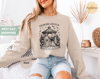 May include: Sand-colored sweatshirt with a graphic featuring two ghosts in a gazebo, pumpkins, and bats. Text reads "GILMORE GHOULS" and "WHERE YOU HAUNT, I WILL FOLLOW". The sweatshirt is a comfortable, casual style.