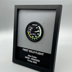 May include: Framed artwork featuring an airspeed gauge and the text "FIRST SOLO FLIGHT." The black frame surrounds a gray background with the gauge at the top and a black panel with text at the bottom. The text includes a name, airport codes, and a date.