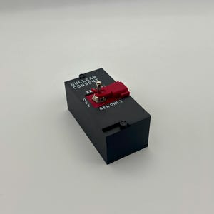 以下が含まれることがあります： 「NUCLEAR CONSENT」と「REL ONLY」の文字が入った、赤いトグルスイッチが付いた黒い長方形の箱。 スイッチは「OFF」の位置にあります。 箱にはシルバーのトグルスイッチと赤いプラスチック製のハウジングが付いています。