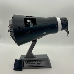 May include: A black and gray model of the Mercury-Redstone 3 Freedom 7 space capsule, mounted on a gray stand. The stand has a plaque that reads "FIRST AMERICAN IN SPACE" and details about the mission. A detached black piece sits to the left.