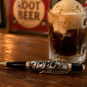 May include: A ballpoint pen with a black and white marbled barrel, gold-toned metal accents, and a black cap and tip. The pen is positioned horizontally in front of a glass mug filled with a dark carbonated beverage and a scoop of vanilla ice cream.