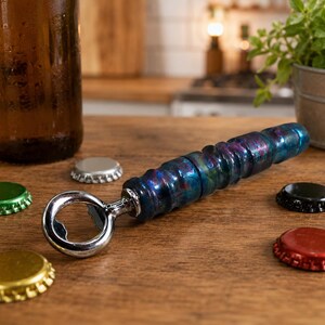 May include: A bottle opener with a cylindrical handle featuring a swirling, marbled pattern of deep blue, purple, and red tones. The opener has a polished chrome metal head and is surrounded by several loose, colourful metal bottle caps.