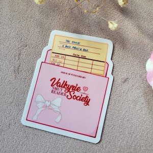 May include: A pink and white sticker with a library card pocket. The card has the title "The House" and "A Book About A Book" written on it. The pocket has the text "Valkyrie Smut Reader Society" in red and white.