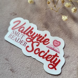 May include: A white sticker with red text that reads "Valkyrie Society" and "Smut Reader". A red heart with the text "VSRS" is on top of a book.
