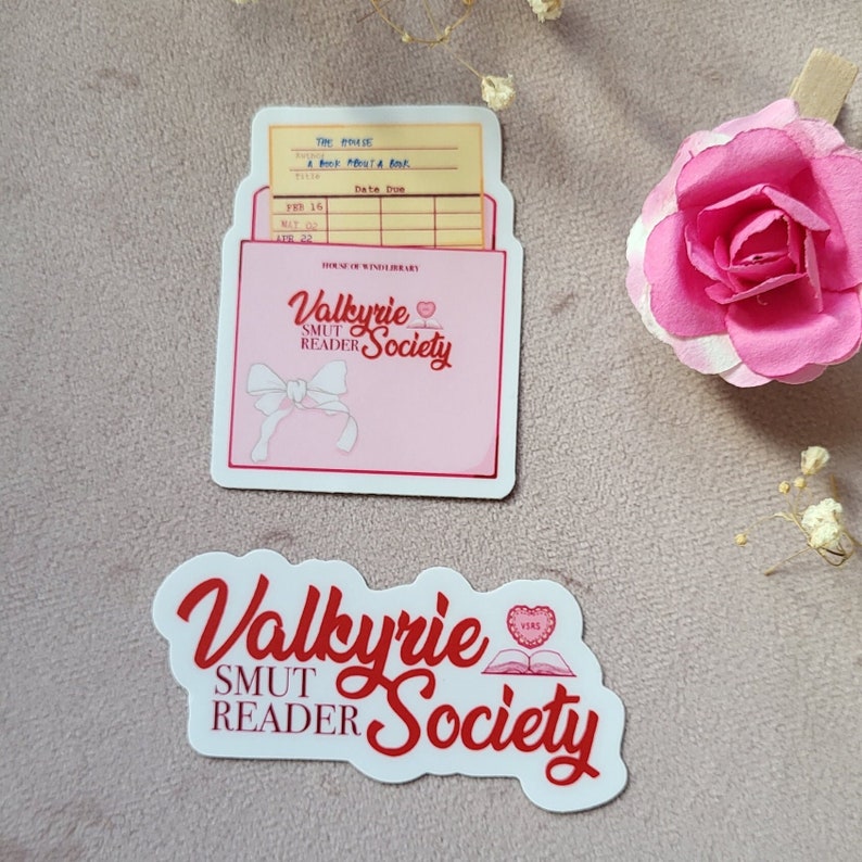 May include: Two stickers featuring the text "Valkyrie Smut Reader Society" in red and white lettering. The first sticker is a pink envelope with a white bow and a glass of wine. The second sticker is a white circle with a red heart and a book.