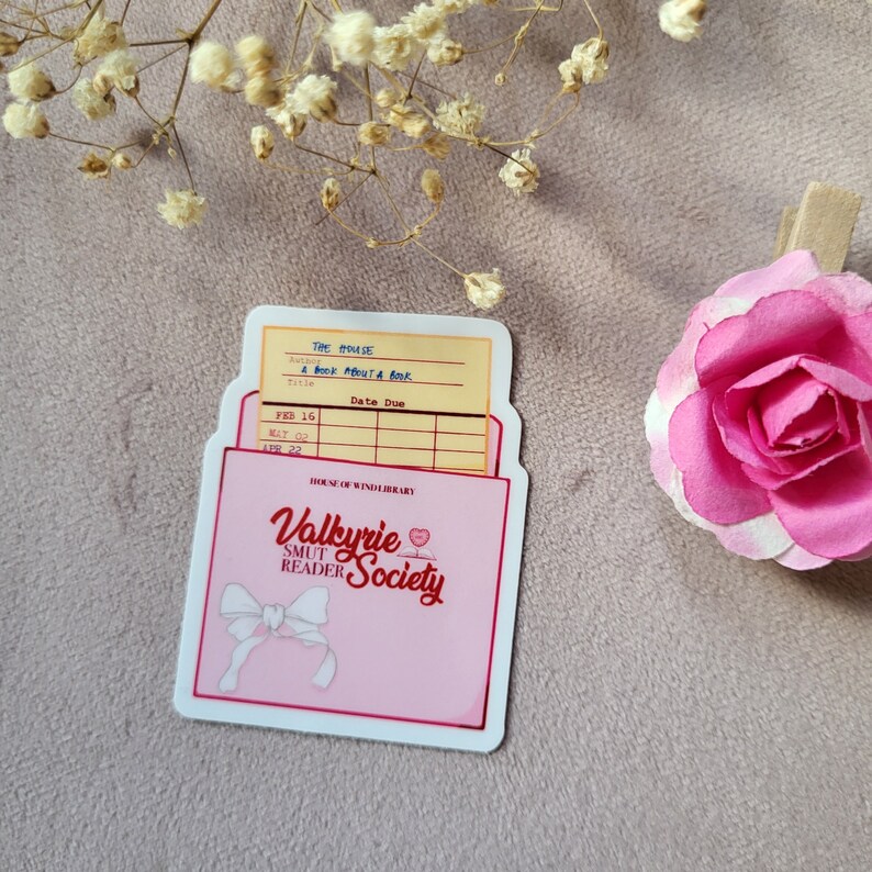 May include: A pink and white sticker with a library card pocket design. The card pocket has a yellow label with the text "The House" and "A Book About A Book". The bottom of the sticker has a pink background with the text "House of Wind Library" and "Valkyrie Smut Reader Society" in red and white text.