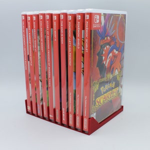 May include: A red and white Nintendo Switch game case stand with 11 video game cartridges inserted. The cartridges include titles like "Tales of Vesperia", "Mario + Rabbids Kingdom Battle", "Shin Megami Tensei V", "Collection of Mana", "Story of Seasons: Friends of Mineral Town", "Mega Man 11", "The End is Nigh", "Pokemon: Let's Go Pikachu", "Pokemon Shield", and "Pokemon Scarlet".