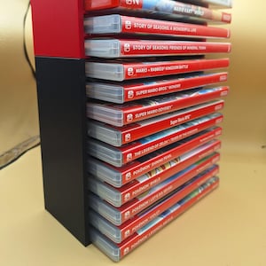 May include: A black and red game cartridge holder filled with Nintendo Switch game cases. The cases are red with white text and feature game titles like "Super Mario Odyssey" and "Pokémon Shield."