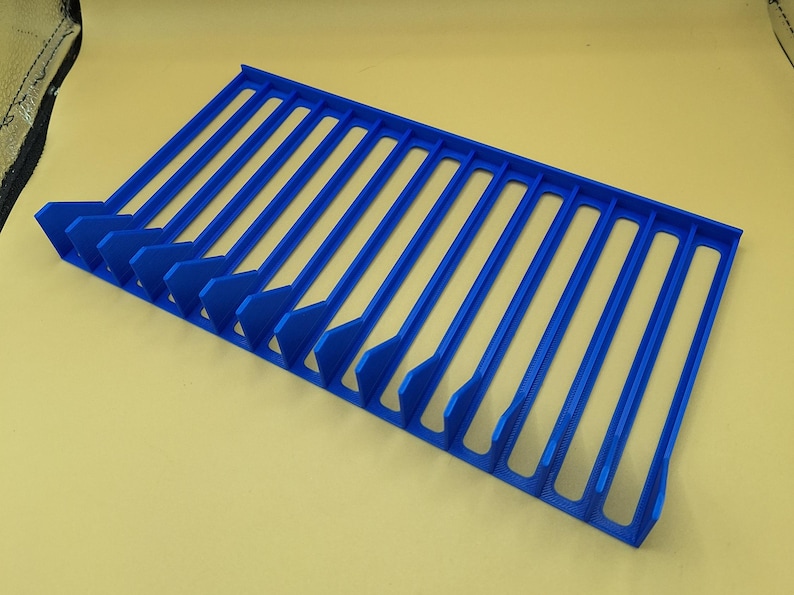 Diagonal right view from above of a blue video game or DVD storage tray in 15 slots featuring wall extensions at the front of each slot to better secure the cases and a slightly elevated back wall. Each slot has a large hole in the base.