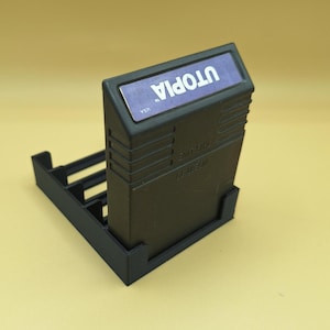 May include: A black plastic stand for a video game cartridge. The cartridge is labeled "Utopia" and "USA".