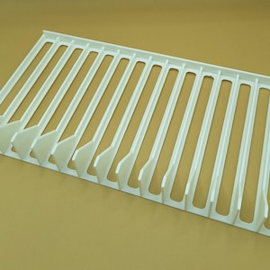 Diagonal right view from above of a white video game or DVD storage tray in 15 slots featuring wall extensions at the front of each slot to better secure the cases and a slightly elevated back wall. Each slot has a large hole in the base.