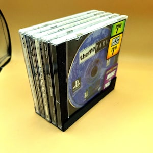 May include: A stack of six PlayStation 1 video game cases, including "Theme Park", "Mamma Mia! The Musical", "DivO", "Ancora", "Saturday Night", and "Arista". The cases are arranged vertically in a black plastic holder. The "Theme Park" case has a blue and purple disc with the game title in white text.