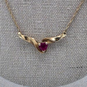 May include: A gold-toned necklace with a heart-shaped, deep red gemstone pendant. The pendant is set in a swirling, stylized design. The chain is delicate and appears to be a cable link style. The necklace is displayed on a textured, neutral-colored surface.