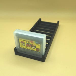 May include: A black plastic game cartridge holder with a single game cartridge in a clear plastic case. The game cartridge has Japanese text and colorful graphics. The holder has multiple slots for additional game cartridges.