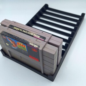 May include: Two gray Super Nintendo game cartridges are stacked vertically in a black plastic game cartridge holder. The game cartridges are for the game "Bass Masters Classic".