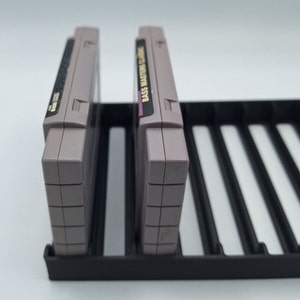 May include: Two gray Super Nintendo Entertainment System game cartridges are displayed in a black plastic storage case. The cartridge on the left has a white label with black text. The cartridge on the right has a white label with black text that reads "Bass Master Classic".