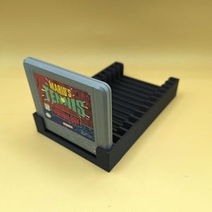 May include: A black plastic stand with slots to hold multiple Virtual Boy game cartridges. A gray Virtual Boy game cartridge is partially inserted into the stand. The game cartridge has the title "Mario's Tennis" printed on it.