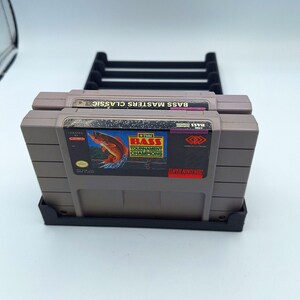 May include: Two gray Super Nintendo game cartridges in a black plastic stand. The game cartridge on top has the text "Bass Masters Classic" and the game cartridge on the bottom has the text "1991 Bass Tournament Champions" and a picture of a fish.