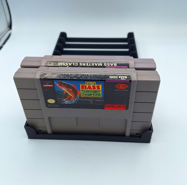 May include: Two gray Super Nintendo game cartridges in a black plastic stand. The game cartridge on top has the text "Bass Masters Classic" and the game cartridge on the bottom has the text "1991 Bass Tournament Champions" and a picture of a fish.