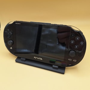 May include: A black PlayStation Vita handheld gaming console with a black stand. The console has a black screen with the text "PS VITA" in gold lettering. The console has buttons on the right side, including "Select", "Start", and directional buttons.