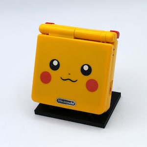 May include: A yellow Game Boy Advance SP with a Pikachu design. The console has a red Nintendo logo and a black base.