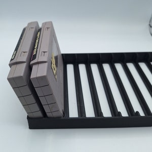 May include: Two gray Nintendo Entertainment System game cartridges are stacked vertically in a black plastic game cartridge holder. The game cartridges are labeled "Bass Masters Classic" and "WWF Wrestlemania".