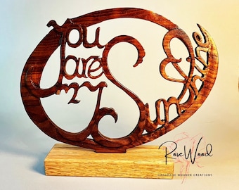 Può includere: Un cartello in legno con le parole "You are my sunshine" intagliate su di esso. Il cartello è in palissandro e ha una finitura in legno naturale. È montato su una piccola base in legno.
