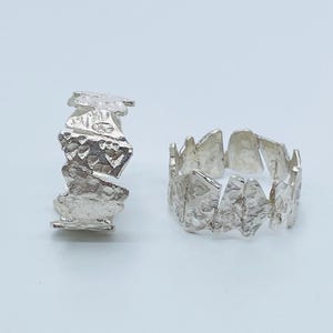 May include: Two silver rings with a highly textured, irregular surface. One ring is positioned vertically on its side, while the other is lying flat, displaying a circular band composed of multiple jagged, metallic segments with a matte finish.