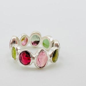 Op de afbeelding: Een zilveren ring met een reeks ovale edelstenen in verschillende kleuren. De edelstenen hebben roze, groene en rode tinten. De ring wordt getoond tegen een witte achtergrond.