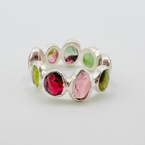 May include: A silver ring with a repeating pattern of oval gemstones in various colours. The gemstones include pink, red, green, and yellow hues. The ring is displayed on a white backdrop.