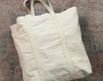 Op de afbeelding: Een grote, off-white canvas tote bag met dikke, witte banden. De tas is gemaakt van een zachte, textuurrijke stof en heeft een ruim interieur.