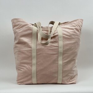 Könnte beinhalten: Eine große, rosafarbene Tragetasche mit dicken, cremefarbenen Henkeln. Die Tasche ist aus einem weichen, samtigen Material.