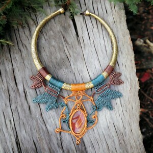 Könnte beinhalten: Eine goldene Choker-Kette mit einem großen Anhänger aus orangefarbenem Achat und komplizierten Knotendetails in den Farben Türkis, Braun und Orange.