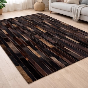 Może przedstawiać: Prostokątny dywan z patchworkowym wzorem. Dywan ma mieszankę ciemnobrązowych, czarnych i beżowych prostokątnych elementów ułożonych w liniowy wzór. Dywan leży na jasnej drewnianej podłodze.