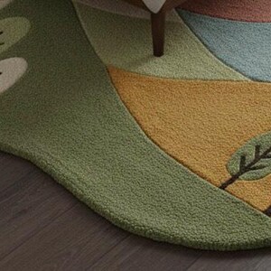May include: A whimsical, landscape-themed rug with a unique, irregular shape. The rug features a design of rolling hills in shades of green, yellow, and blue, with stylized trees. The rug is placed on a dark wood floor.