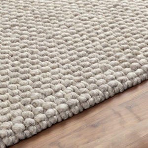 May include: Close-up of a neutral-toned, textured rug with a woven, nubby design. The rug's surface is a light beige colour, and the texture is created by the raised, rounded loops of the weave. The rug is placed on a wooden floor.