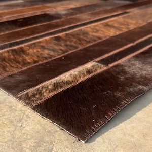 May include: A rectangular rug with a patchwork design. The rug features various shades of brown, from light tan to dark chocolate, creating a textured appearance. The rug is made of a natural material, likely animal hide, and has a soft, plush texture.