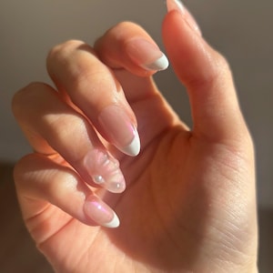 Peut inclure: Gros plan d'une main avec plusieurs ongles peints avec une french manucure à pointe blanche. Certains ongles ont de petits embellissements perlés.