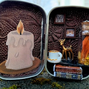 May include: An open, silver-colored tin with a miniature scene. One side features a candle with a flame, the other a deer figurine, tiny framed pictures, a teacup, a glass jar, and miniature books. The background is a textured brown.