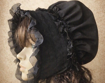 May include: A black bonnet featuring a gathered crown and a wide, ruffled sheer trim along the front edge. The headwear is constructed from a solid black fabric with a soft texture, accented by a matching black ribbon tie under the chin area.