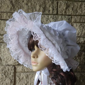 May include: A white bonnet with a wide brim, trimmed with delicate lace and a pale pink lining. The bonnet features a ruffled design and is tied with a white ribbon. The image shows a close-up of the bonnet on a mannequin.