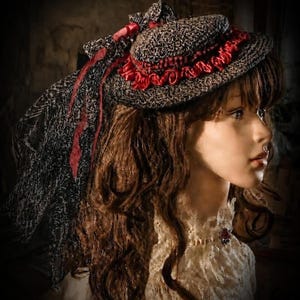 May include: A dark gray and black textured hat featuring a vibrant red ruffled ribbon trim around the crown. A long, sheer black lace veil with a subtle pattern hangs from the back of the hat, accented by thin red ribbons tied in a bow.