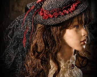 May include: A dark gray and black textured hat featuring a vibrant red ruffled ribbon trim around the crown. A long, sheer black lace veil with a subtle pattern hangs from the back of the hat, accented by thin red ribbons tied in a bow.