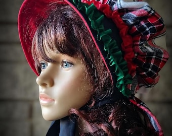 May include: A bonnet featuring a red velvet brim with a green ruffled trim. The crown is made of a red, white, and black plaid fabric with a red ruffled accent. The bonnet includes black fabric ties and is displayed on a mannequin head.