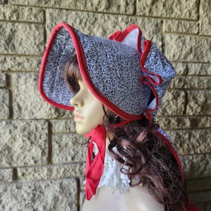 May include: A bonnet with a floral pattern in black and white, trimmed with red fabric. The bonnet is on a mannequin head. The bonnet has a wide brim and a red ribbon tie. The mannequin is wearing a red scarf with a white lace collar.