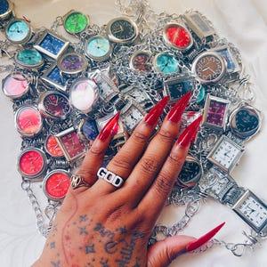 May include: A collection of silver-tone wristwatches with various colored faces, including red, blue, green, and black, linked together by silver chains. The watches feature both round and rectangular cases with clear numerical dials and silver hands.