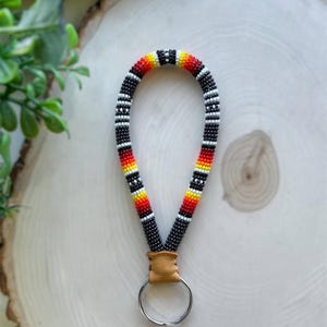 May include: Handmade beaded keychain lanyard with a black, white, red, and yellow pattern. The lanyard is attached to a small piece of tan leather and a silver key ring. The beaded portion is approximately 6 inches long.